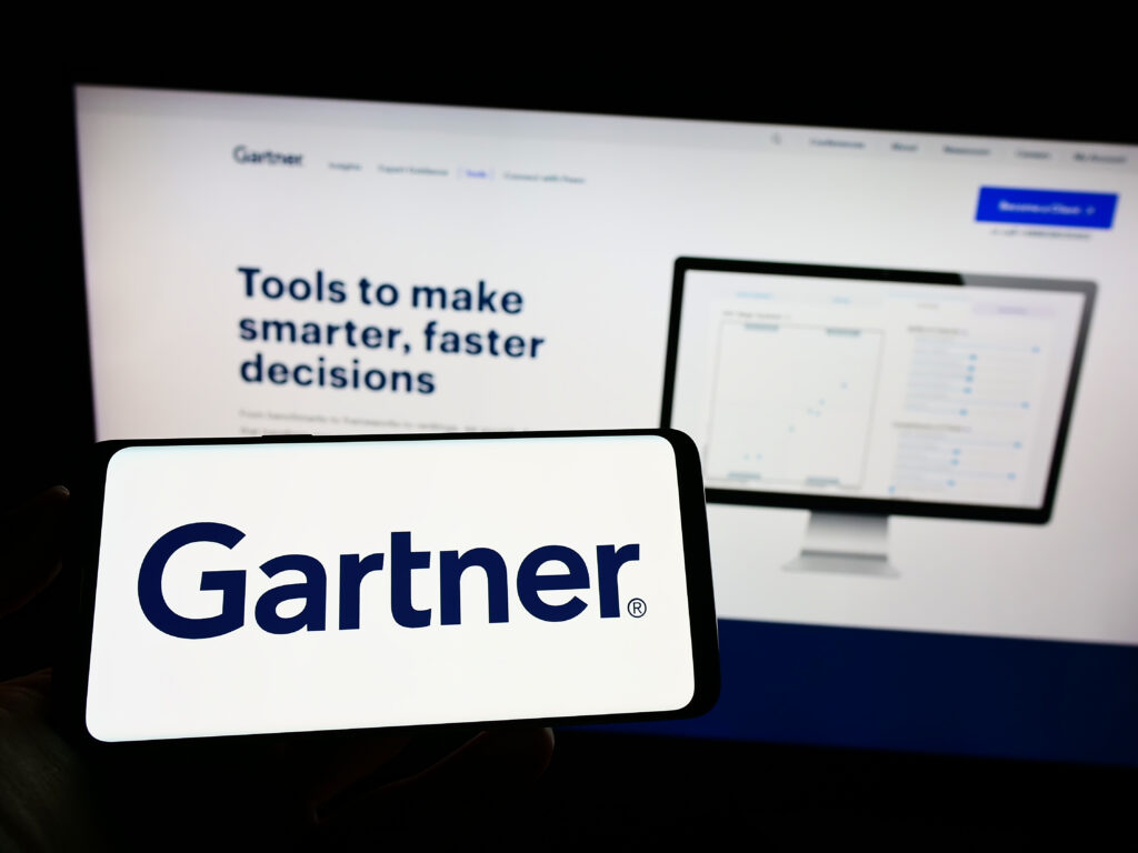 Smartphone displaying Gartner logo in front of a computer screen showcasing Gartner's website with tools for making smarter, faster decisions.