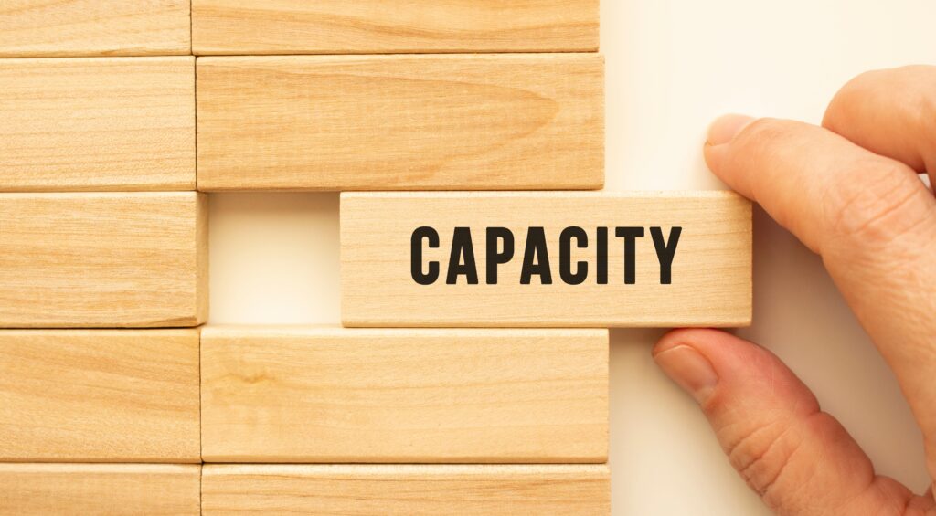 Hand placing a wooden block labeled 'Capacity' into a puzzle, symbolizing the importance of capacity management in completing the structure of effective resource planning and business operations.